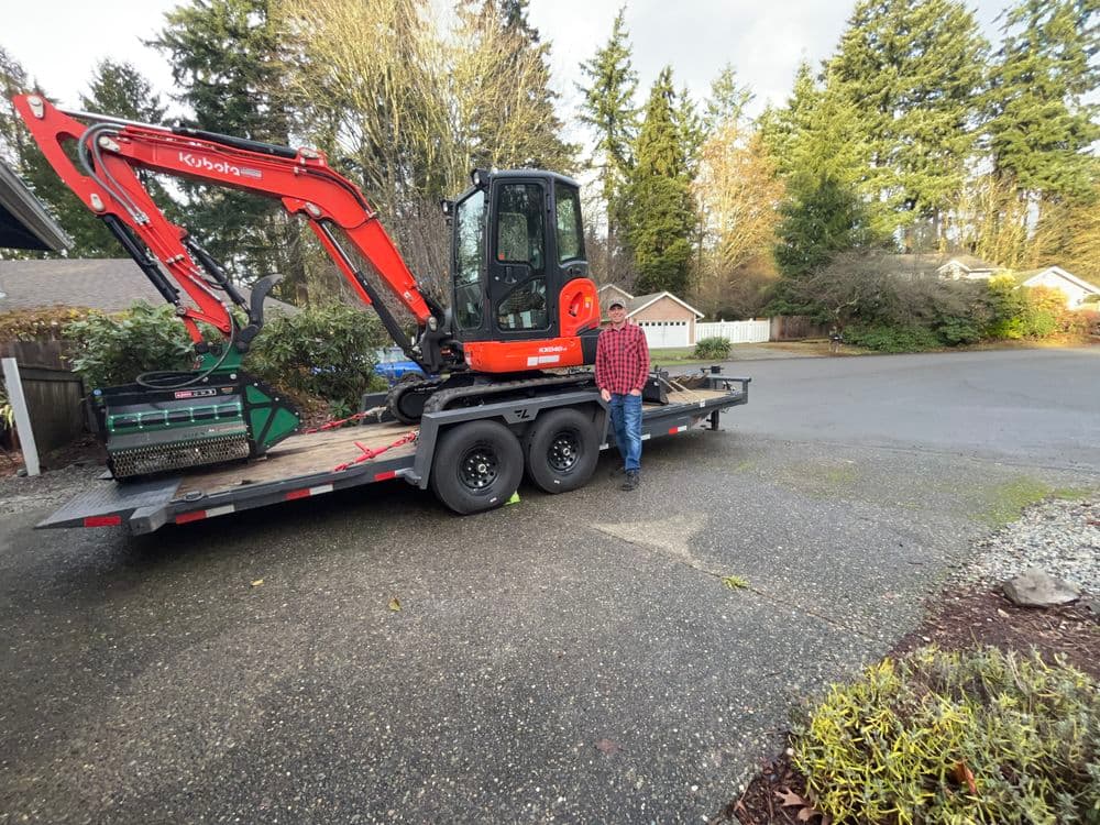 Excavator on a trailer with a person standing beside it in a residential area.
