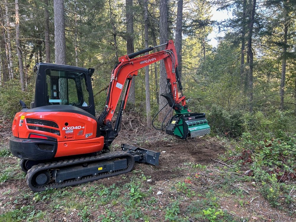 Excavator with forestry attachment in wooded area, ready for land clearing action.