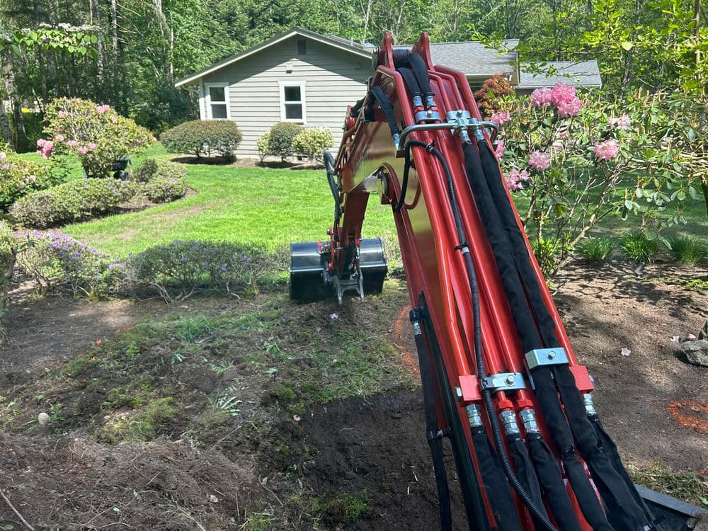 Excavator digging in garden with flowers and house in background on a sunny day.