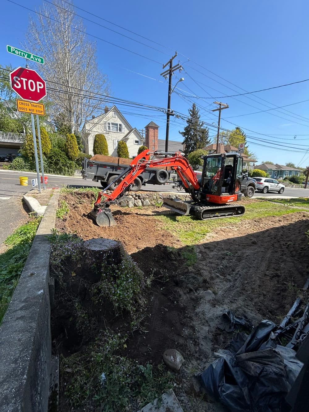 Excavator digging in front yard near stop sign on a sunny day, landscaping work underway.
