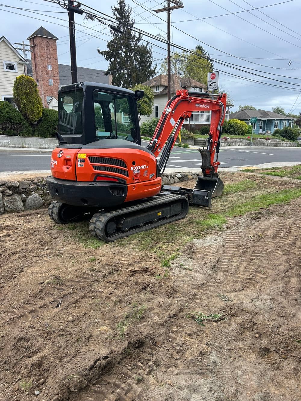 Compact orange excavator parked on a dirt lot near a residential area and power lines.