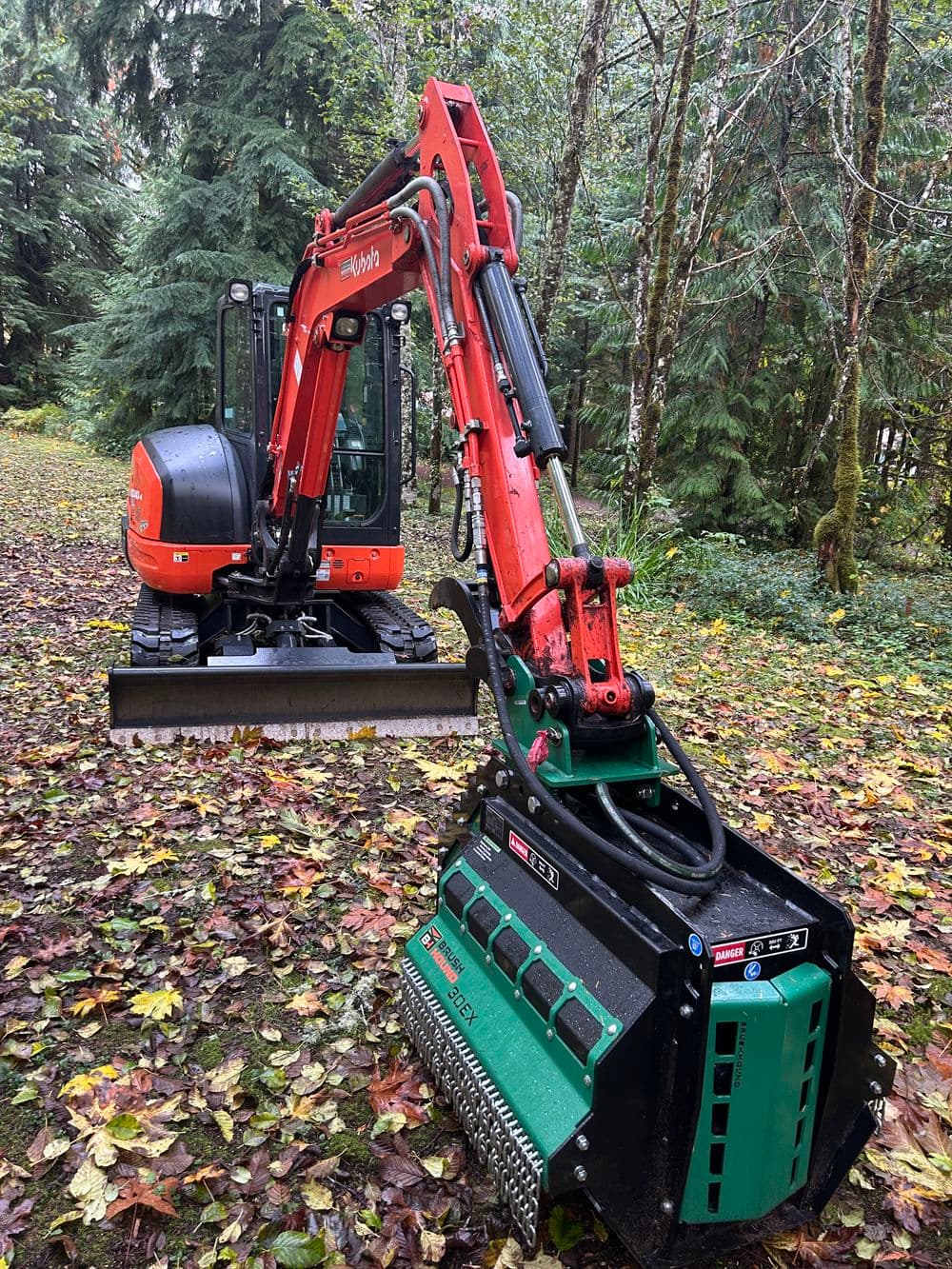 Excavator with a mulching head in forested area surrounded by autumn leaves.